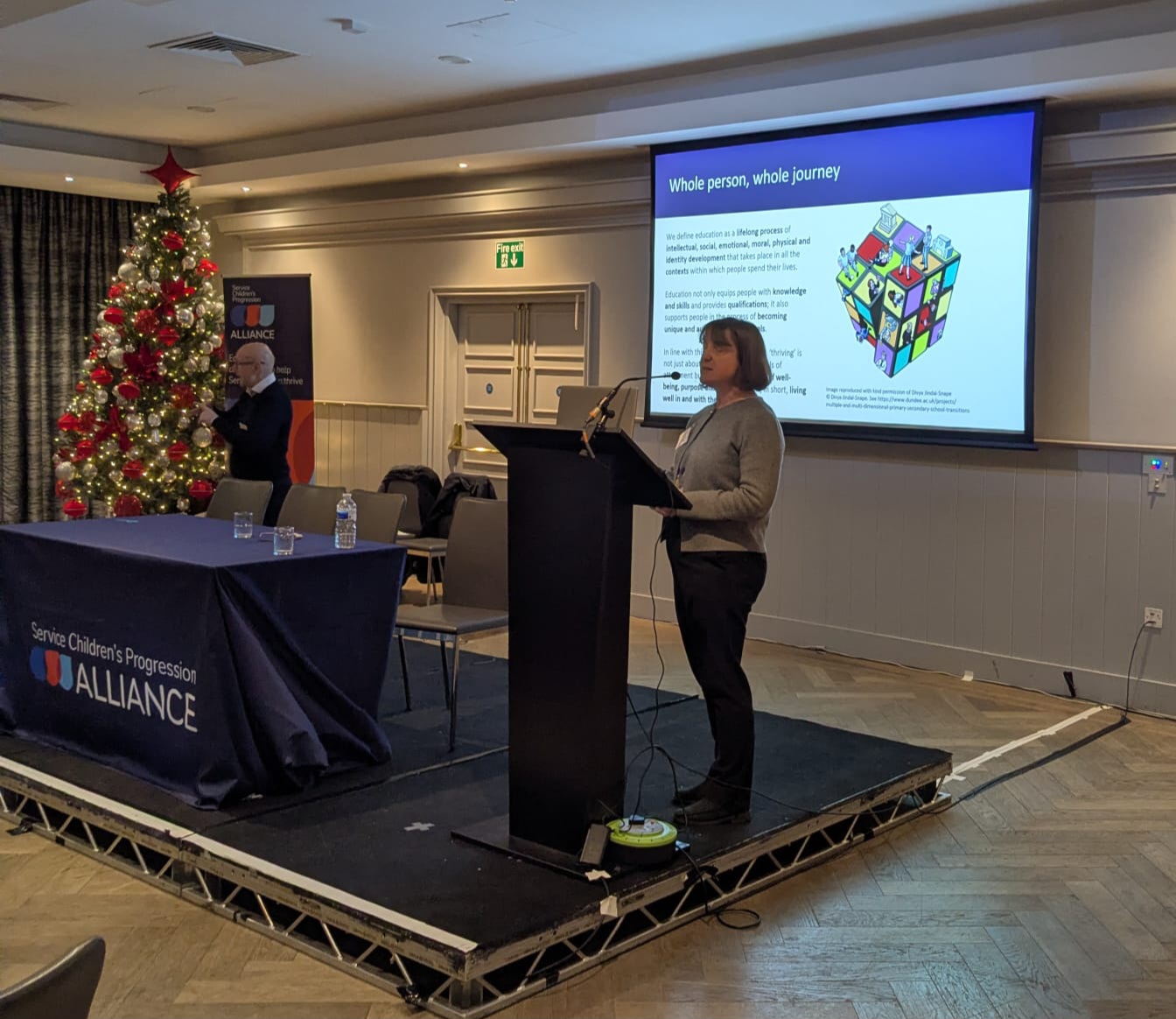 A woman stands at a lectern. Behind her is a screen with an image projected